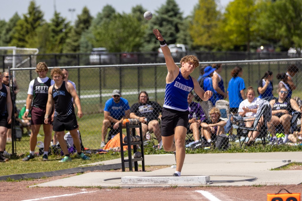 A male athlete in a blue tank top throws a shot put at a track and field event, with spectators seated in the background.