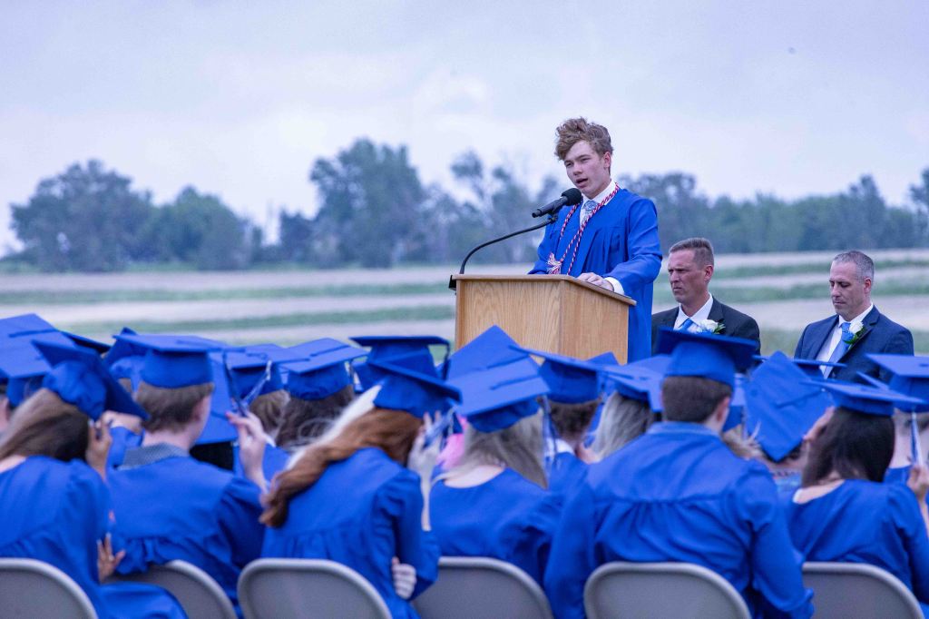 A graduation ceremony scene with a student in a blue cap and gown delivering a speech at a podium, while fellow graduates, also in blue caps and gowns, listen attentively in the foreground. The background shows a green landscape and cloudy sky.