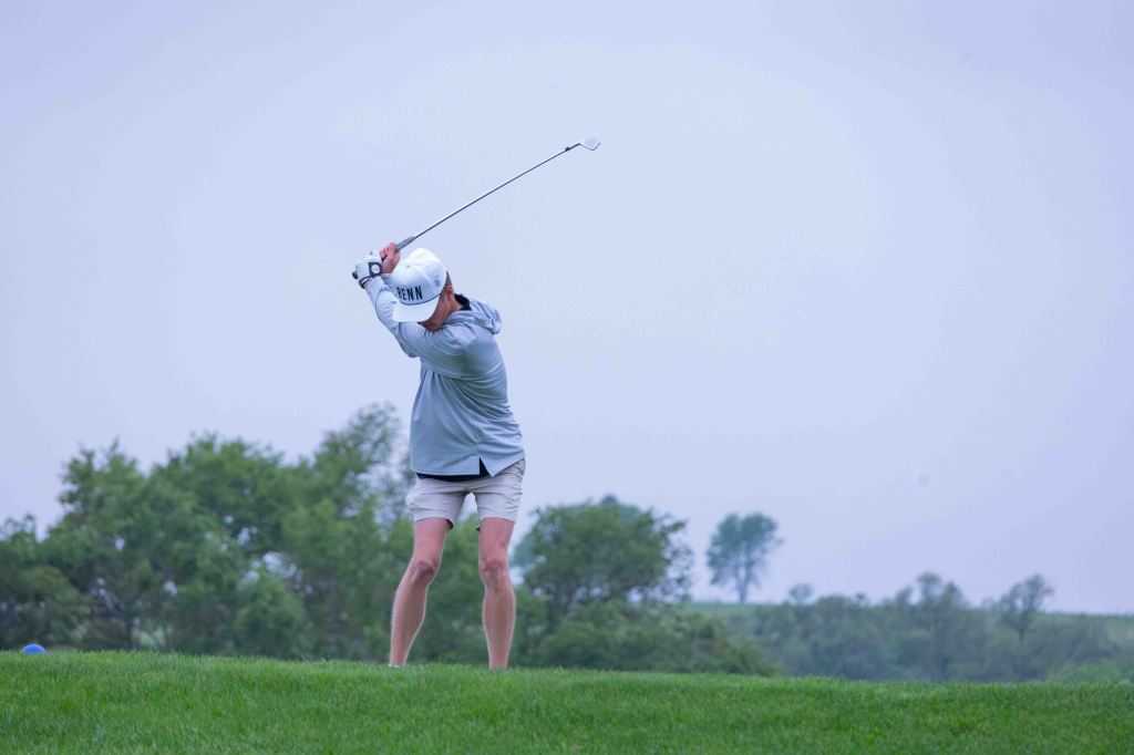 A golfer in a gray shirt and white cap is executing a swing on a green golf course under a cloudy sky.