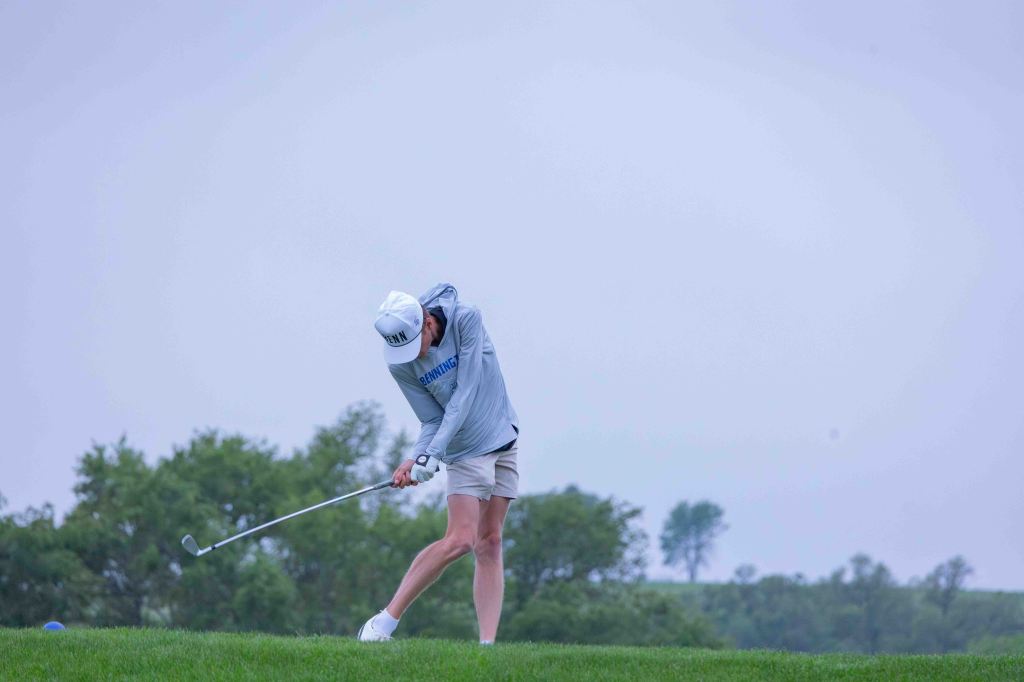 A golfer swinging a club on a grassy field, with a cloudy sky in the background.
