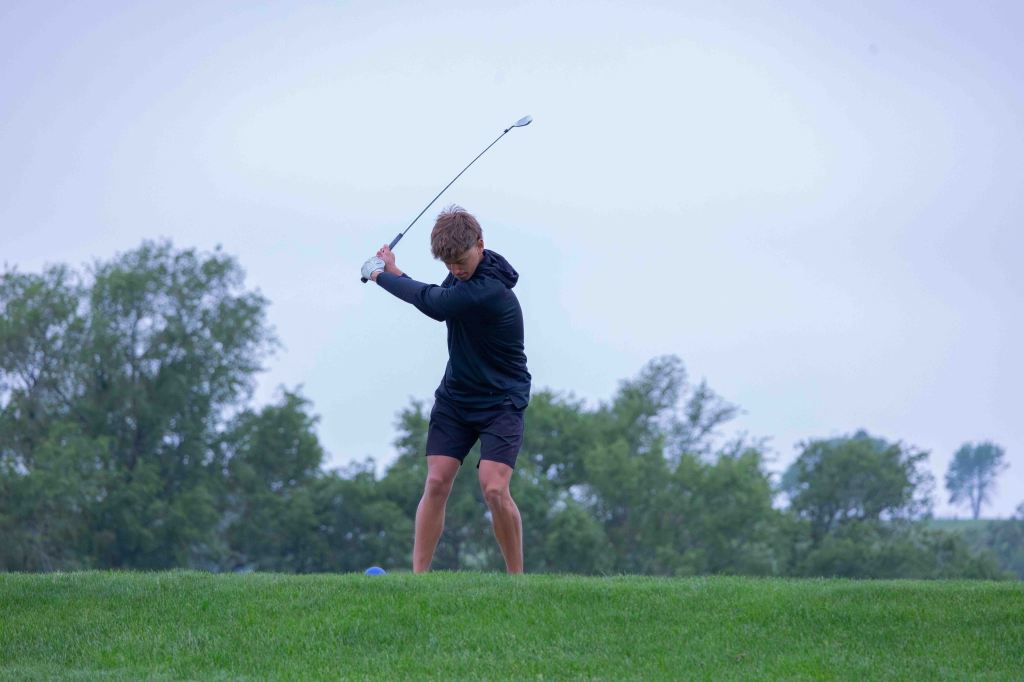 A young golfer in a black hoodie and shorts is swinging a golf club on a grassy course with trees in the background.