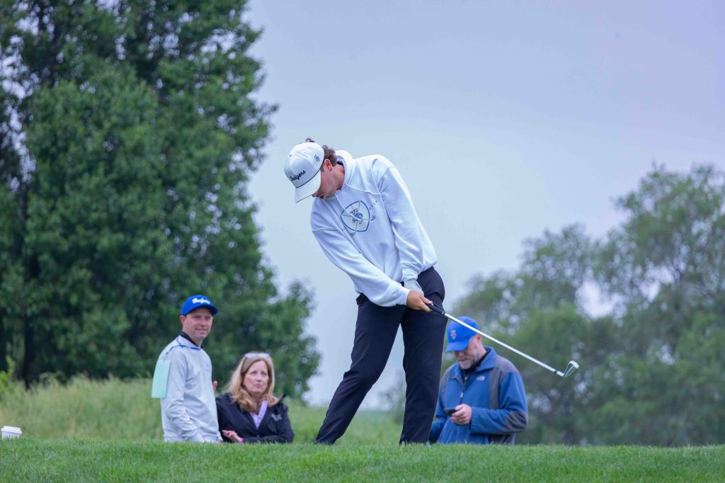 A young golfer prepares to swing a club on a lush green course, with spectators watching in the background.