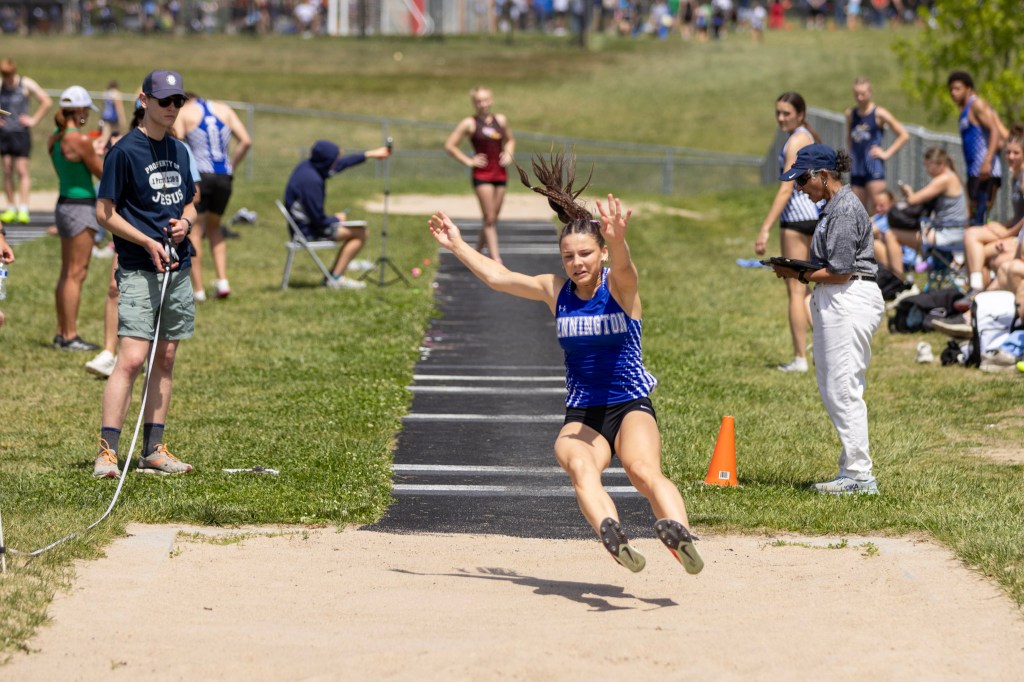 A female athlete in mid-air during a long jump event, wearing a blue and white uniform. Spectators and officials are watching from the sidelines.