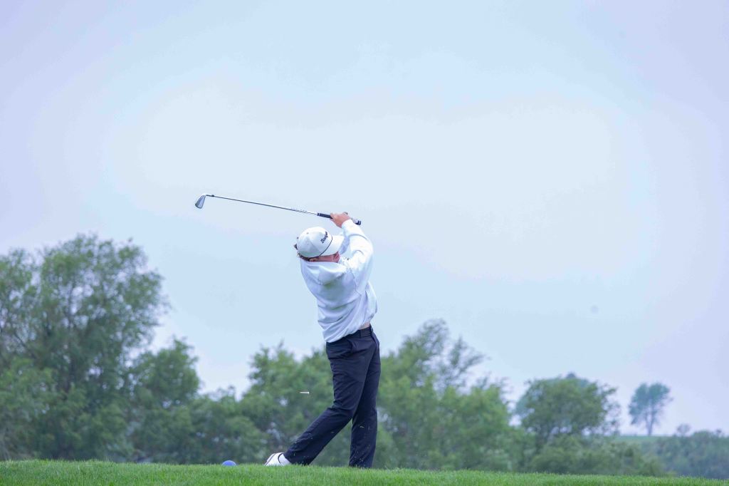 A golfer in a white shirt and hat swings a club on a grassy course, with trees in the background under a cloudy sky.