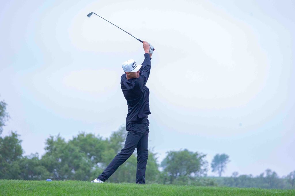 A golfer preparing to swing on a green field, wearing a black outfit and a white cap.