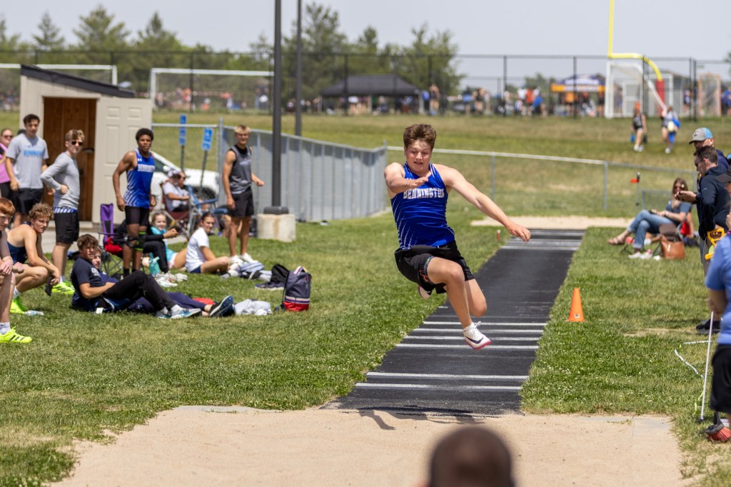 A male athlete in a blue tank top performs a long jump at a track and field event, while spectators cheer from the sidelines.