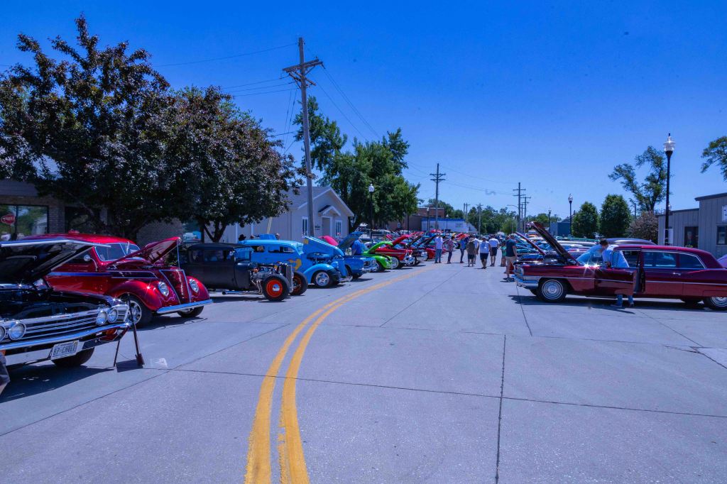 A colorful lineup of classic cars displayed on a sunny street during a car show, with spectators milling about and taking in the vintage vehicles.