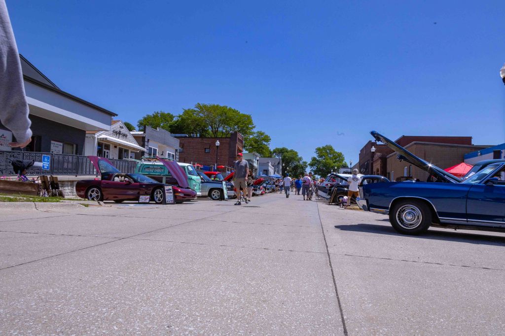 A vibrant street scene featuring various classic cars with hoods raised, alongside crowds of people walking, all under a clear blue sky.