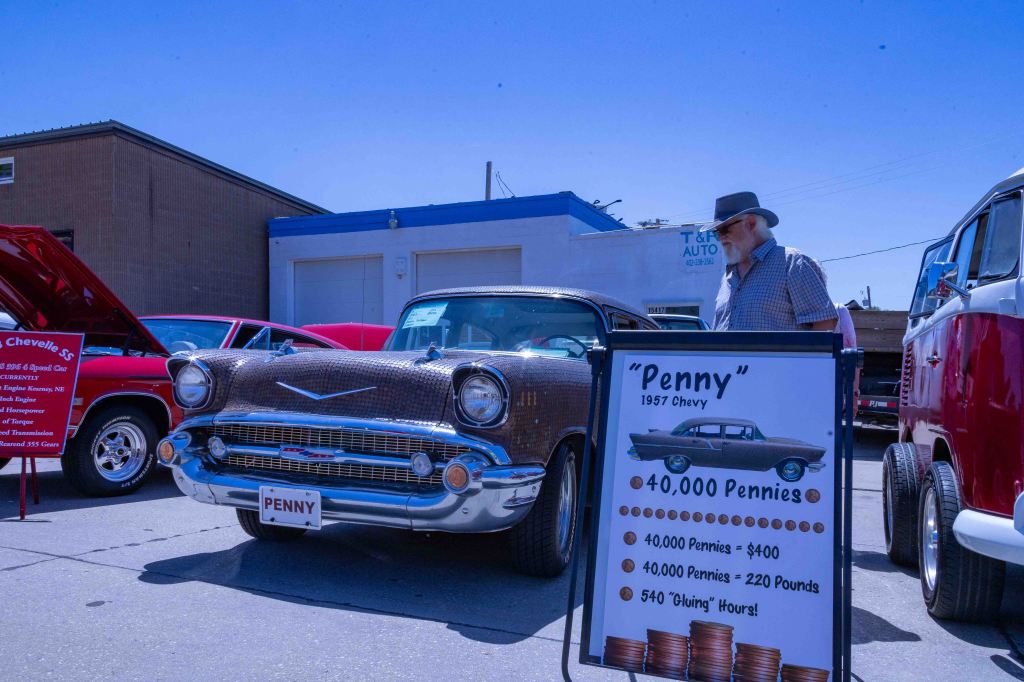 A vintage 1957 Chevy car named 'Penny' displayed at a car show, with a sign detailing its asking price of 40,000 pennies. A man stands next to the car, and other classic cars can be seen in the background under a clear blue sky.