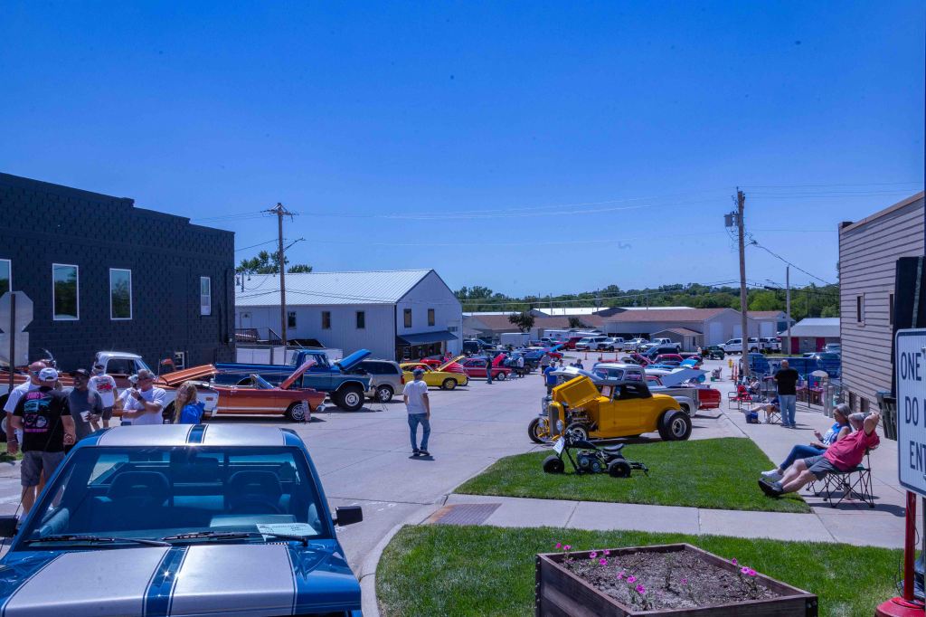 A clear blue sky over a lively street scene featuring a variety of classic cars and trucks, with people gathered around and enjoying the car show.