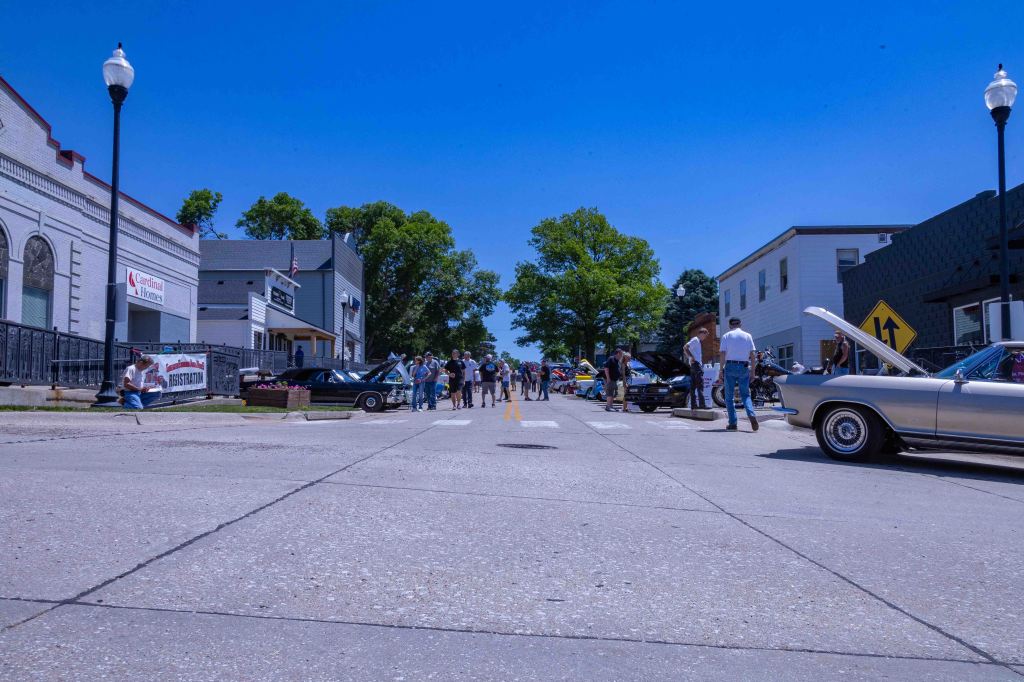 A bustling street scene featuring a classic car show, with attendees admiring various vintage vehicles under a clear blue sky.