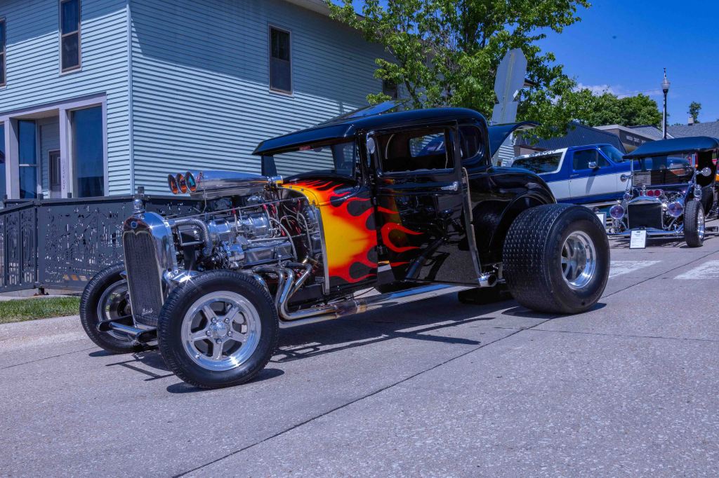 A classic hot rod car with a shiny chrome engine and flame graphic paint job, parked on a street near a residential building, featuring large rear tires and a vintage design.