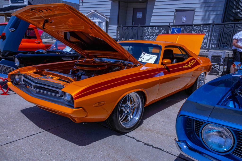 A classic orange muscle car with a striped design, parked with its hood open, showcasing the engine. The scene features other classic cars in the background.