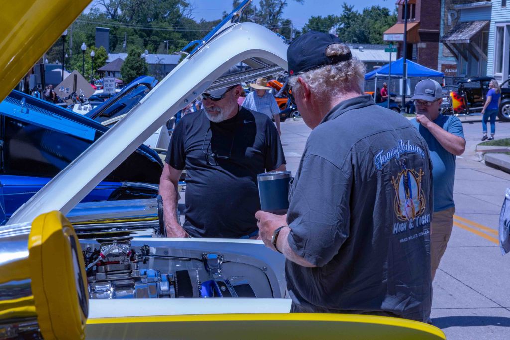 Two men examining the engine of a vintage car at a car show, with colorful cars and onlookers in the background.
