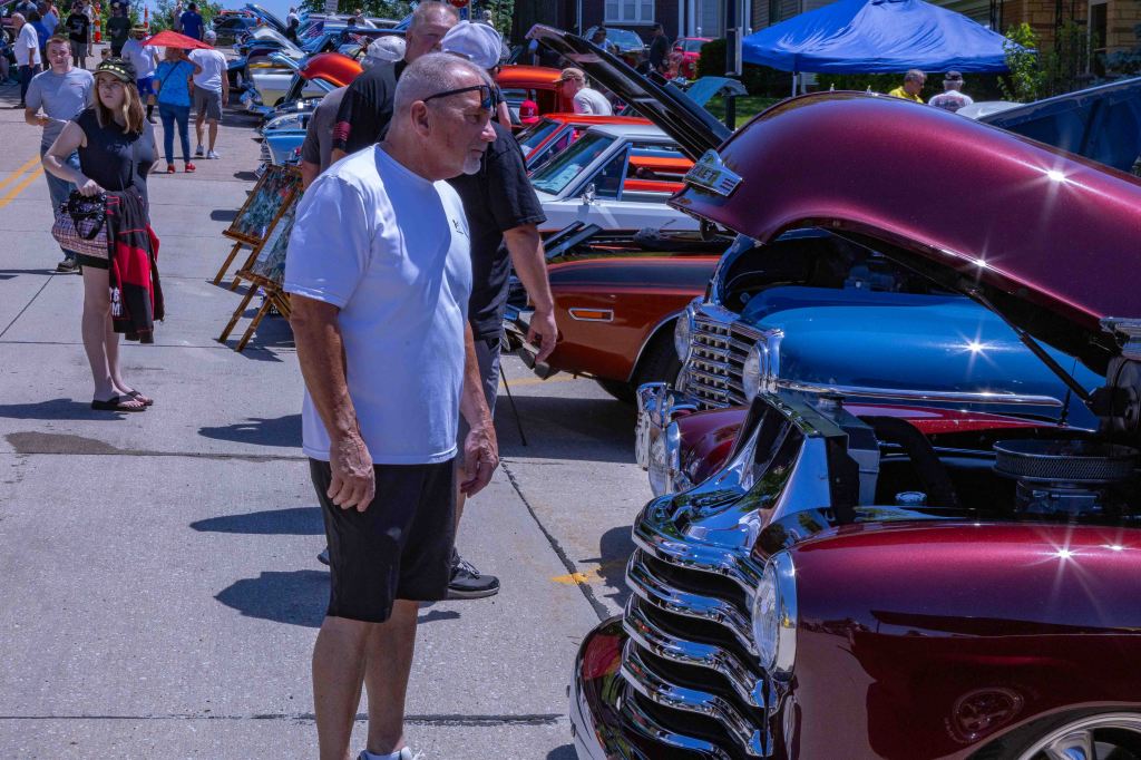 A man in a white shirt examines a classic car with its hood raised at a car show event, surrounded by a crowd and various vintage vehicles under a sunny sky.