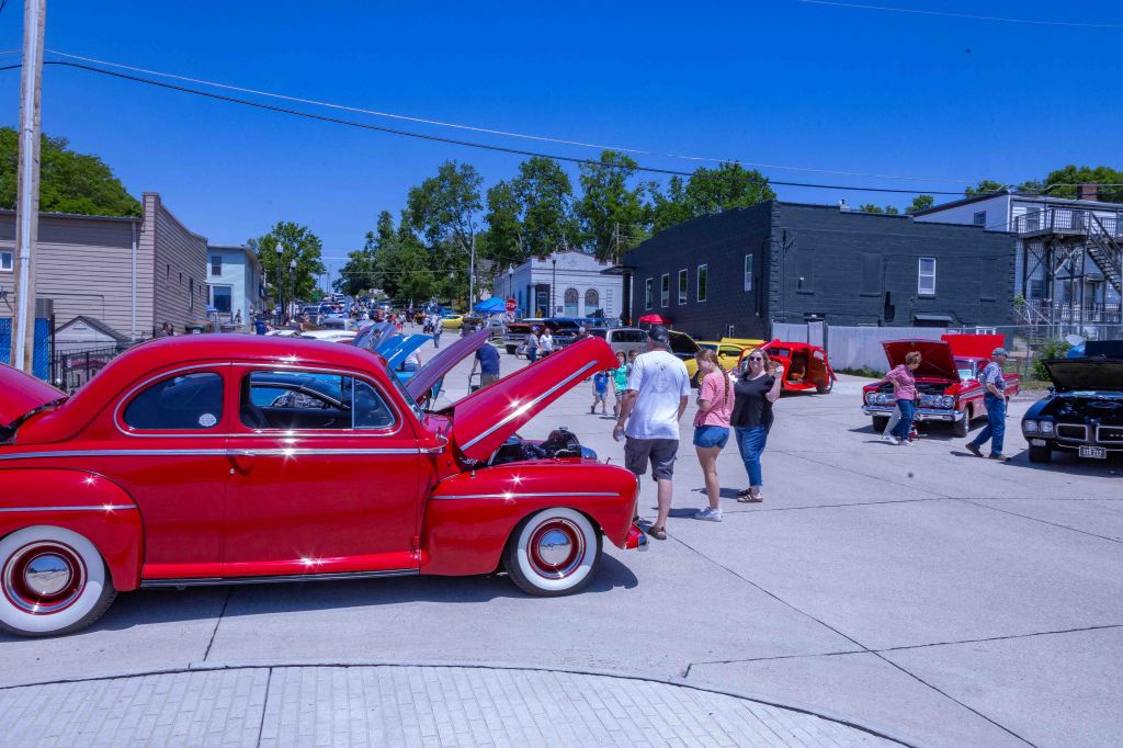 A vintage red car is parked on a sunny street, with several people gathering around other classic cars. The scene is lively, showcasing a car show atmosphere with various car models and attendees enjoying the event.