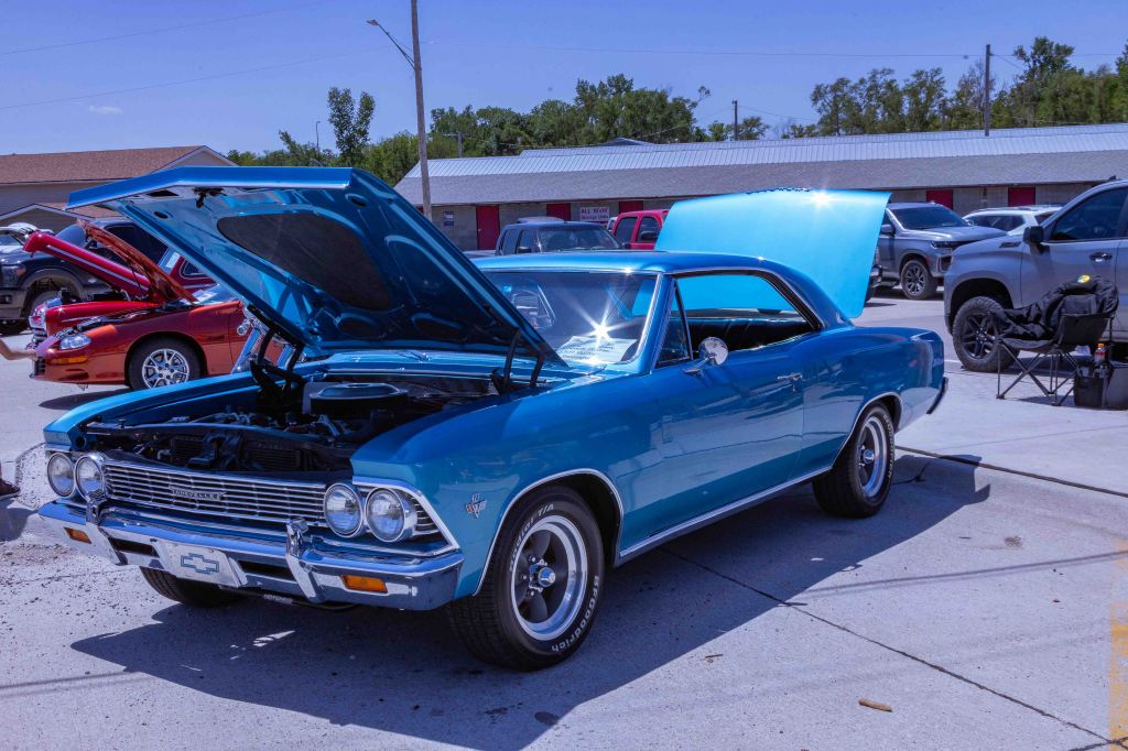 A vintage blue Chevrolet car with the hood open, showcasing the engine, parked at a car show. Other vehicles are visible in the background.
