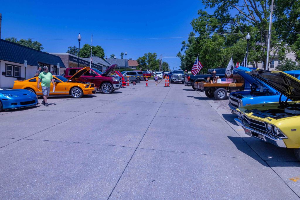 A vibrant car show featuring multiple classic and modern cars lined up on a street, with spectators walking among the vehicles and flags displayed.