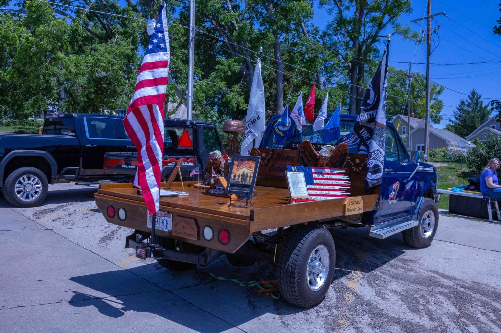 A pickup truck with a wooden bed decorated with various flags, including an American flag, and memorabilia honoring veterans. The truck is parked in a sunny area with trees in the background.