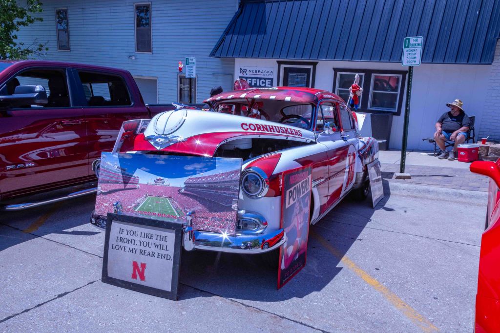 A vintage car decorated in red and white with 'Cornhuskers' lettering, featuring a sign that reads 'If you like the front, you will love my rear end,' alongside a poster of a football stadium. A man sitting in a hat can be seen in the background.