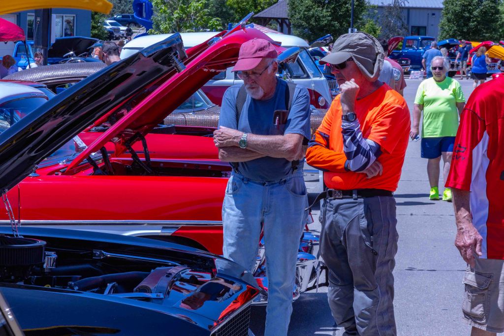 Two men, one wearing a red cap and the other in an orange shirt, are observing a classic car with its hood open at a car show. Brightly colored vintage cars are parked in the background.