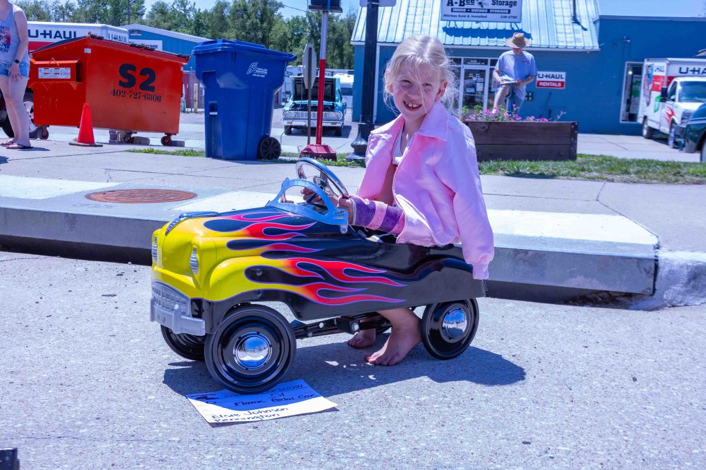 A young girl in a pink jacket is sitting in a small toy car with yellow flames on the side, parked on a sunny street. A sign with her name is placed on the ground next to the car.