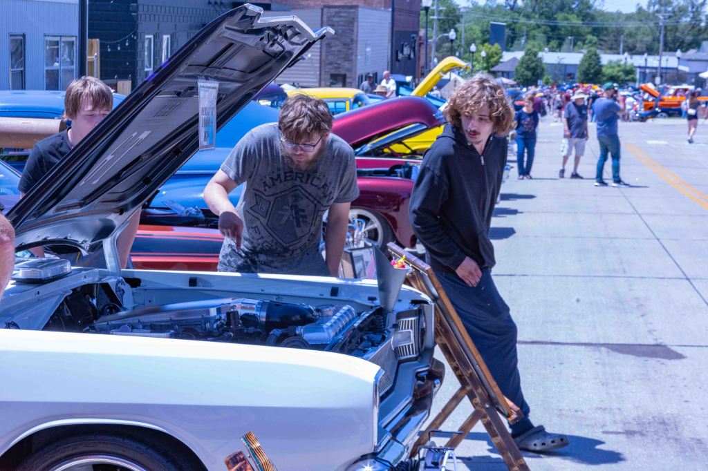 Two young men examining the engine of a classic car at a car show, with various colorful vehicles in the background and an audience observing.