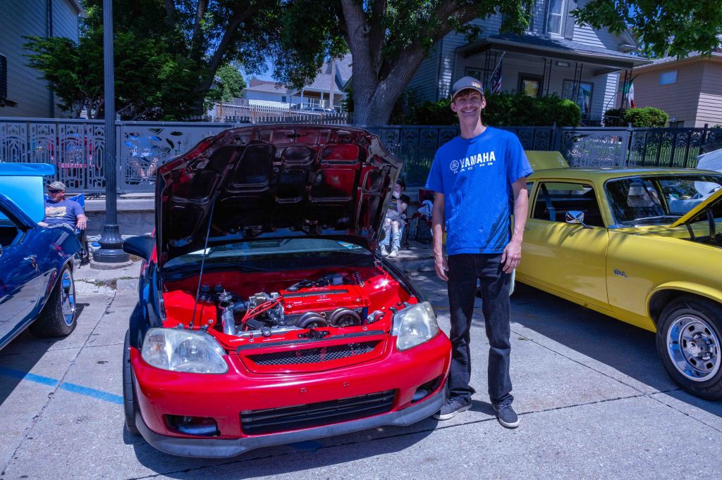 A man in a blue shirt stands next to a red car with its hood open, showcasing the engine. In the background, there are other classic cars and people sitting nearby in a sunny outdoor setting.