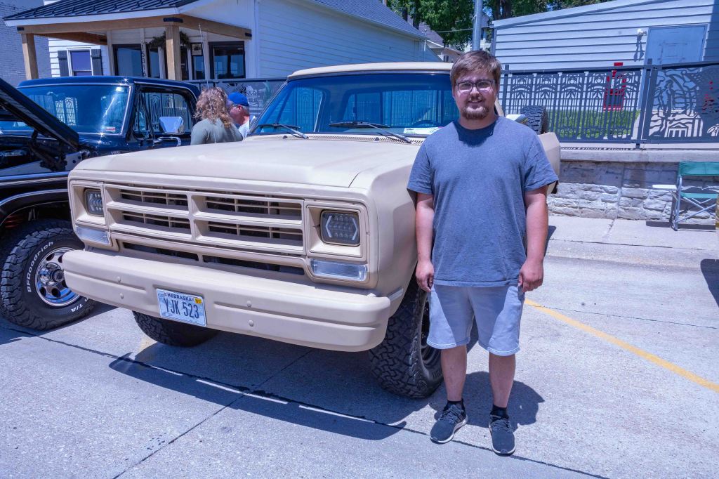 A young man stands next to a vintage beige truck at a car event, with other cars parked in the background.