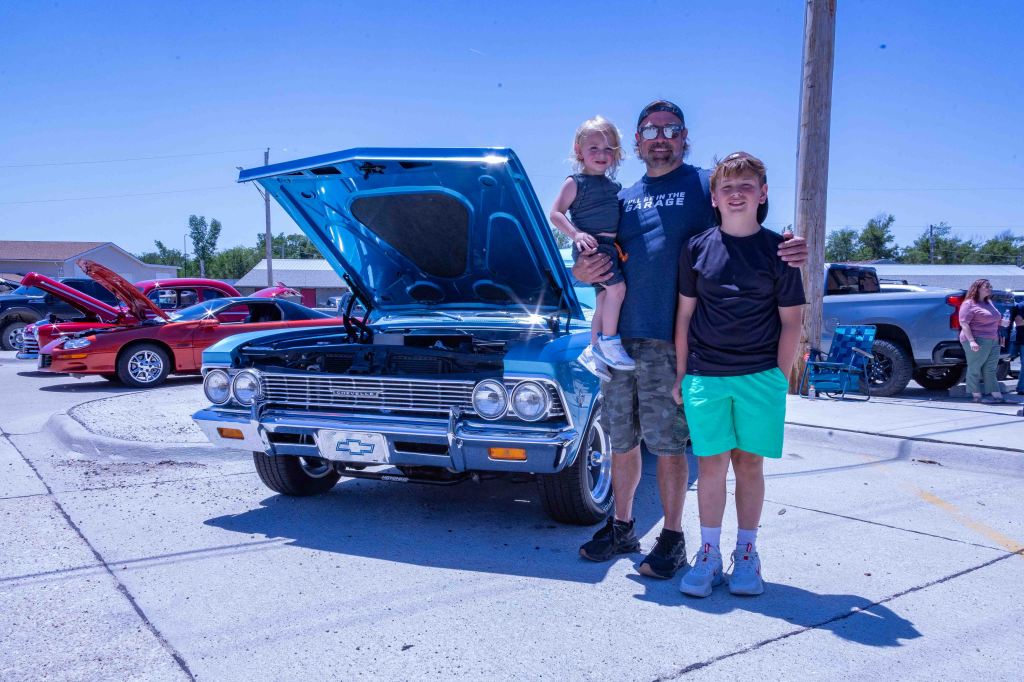 A father stands with his two children, one on his hip, in front of a classic blue Chevrolet car with the hood open. The background shows other vintage cars and people enjoying a car show on a sunny day.