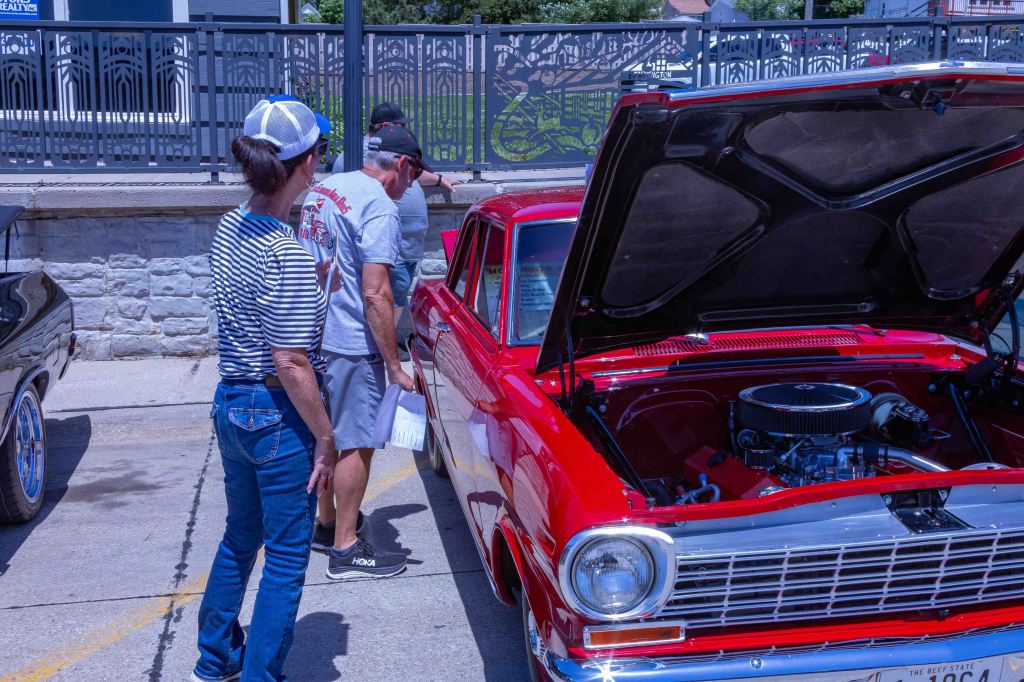 Two people examining a classic red car with its hood open in a parking area, surrounded by a stone wall and a decorative fence.
