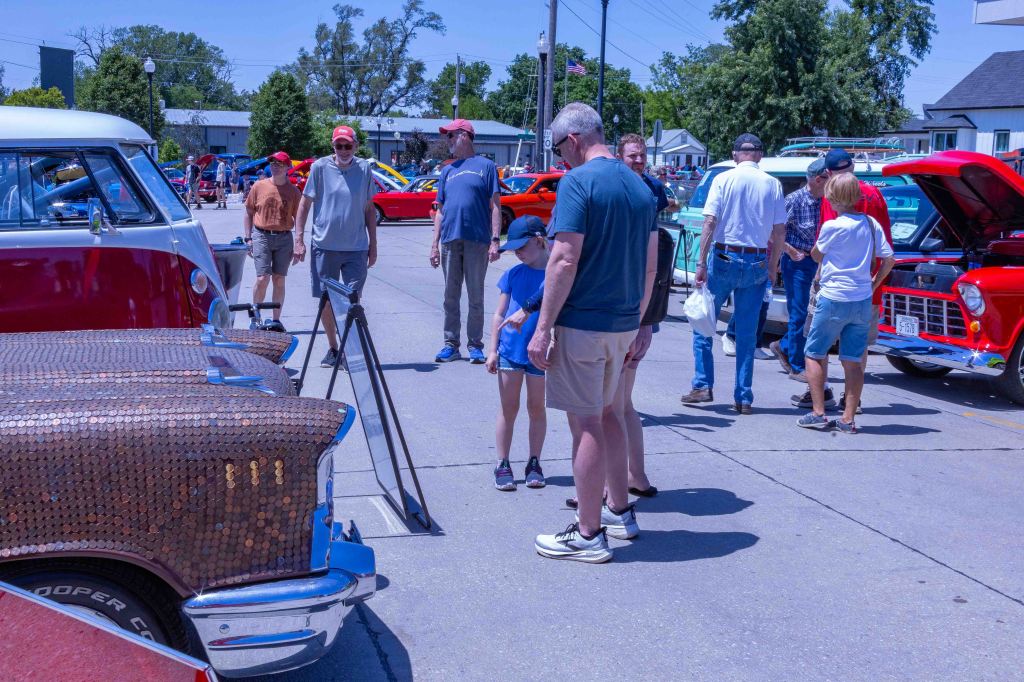 A crowd of people at a car show, with a focus on a vintage car featuring a penny-covered hood. A man and a girl are examining the car, while others are walking around admiring various classic cars in the background.