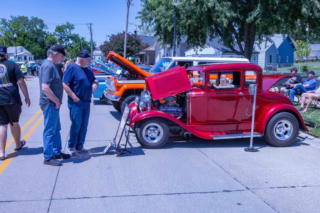 A bright red classic car with its hood open, showcasing the engine, while two men observe it on a sunny day at a car show.