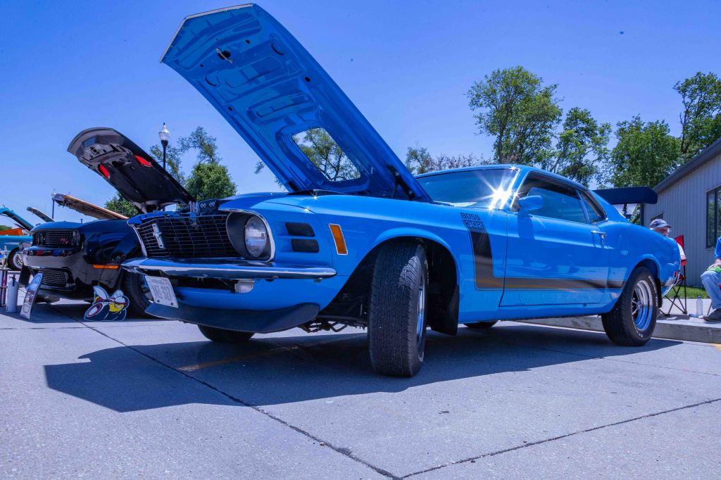 A classic blue muscle car with its hood open, showcasing the engine, parked on a sunny day at a car show. Several other cars and spectators can be seen in the background.