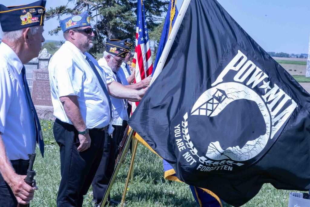 Veterans participating in a memorial ceremony holding a POW-MIA flag, with American flags in the background at a cemetery.