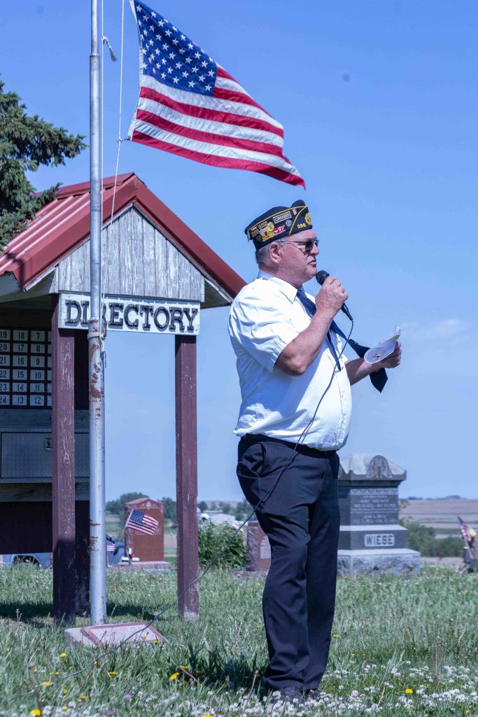 A speaker in a white shirt and veteran's cap addresses an audience while holding a microphone, standing near a flagpole with an American flag. In the background, there are gravestones and a directory sign.