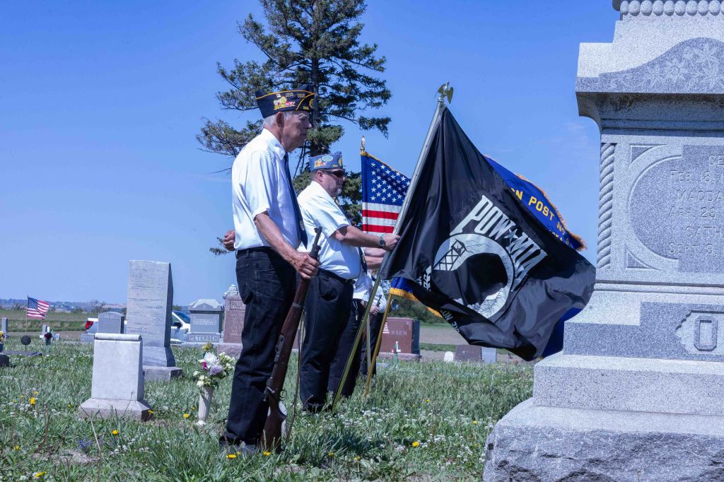 Veterans standing at attention in a cemetery, holding flags, during a commemorative ceremony.