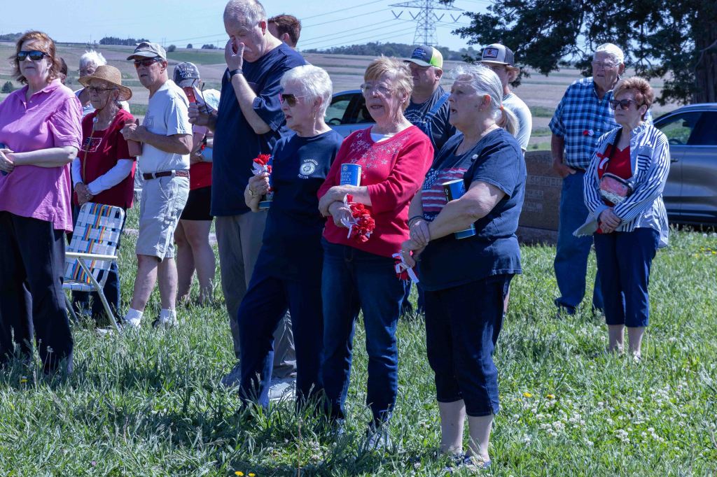 A group of people standing together outdoors, some holding flowers, during a gathering or event in a grassy area. The attendees are dressed in casual clothing, with a mix of colors and patterns, and several individuals are positioned in the background.