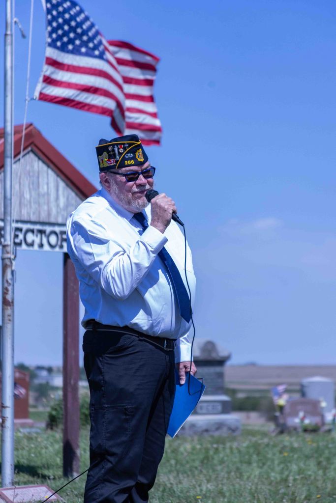 A man in a suit and veterans cap speaking into a microphone, with an American flag waving in the background, at a cemetery.