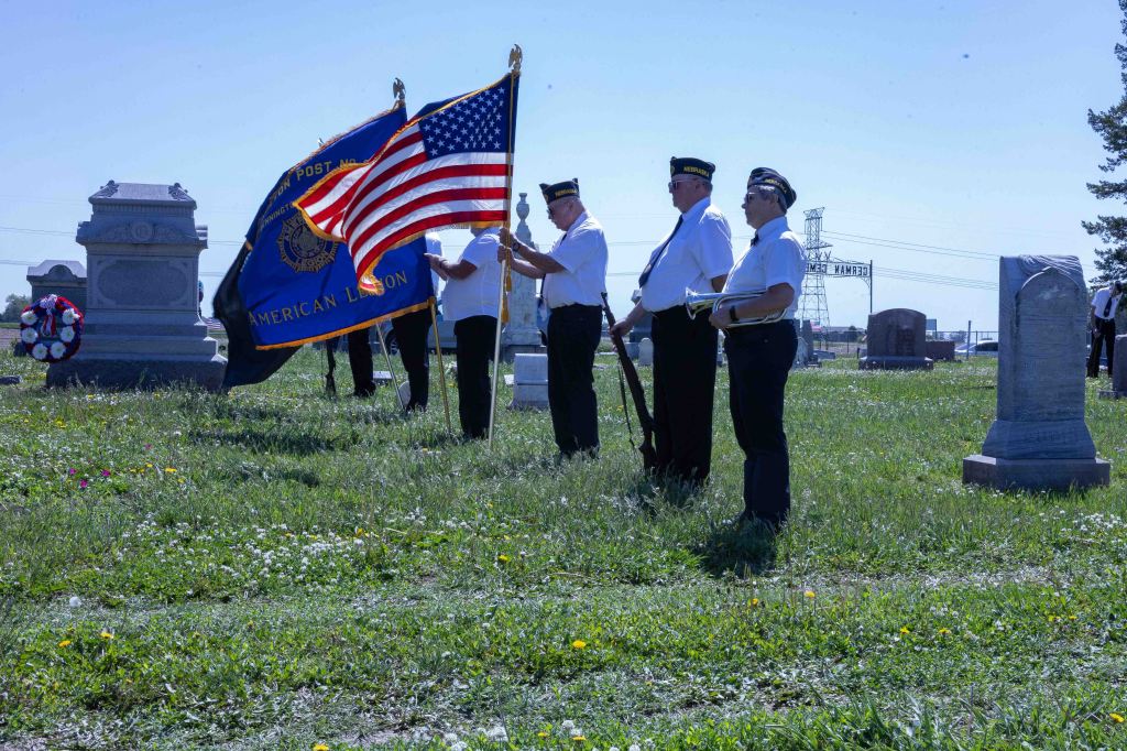 Veterans in uniform holding the American flag and a Post flag while standing in a cemetery during a memorial ceremony.