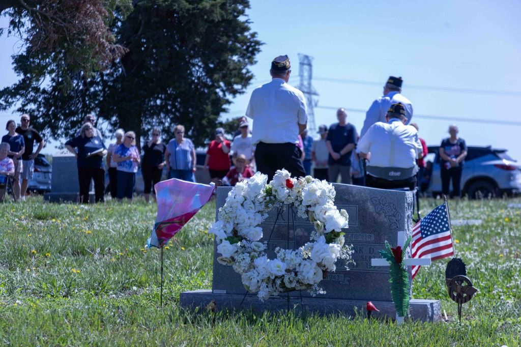 A group of people gathered at a cemetery for a memorial service, with a gravestone decorated with flowers, an American flag, and other tributes in the foreground.
