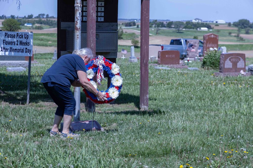 An elderly woman placing a flower wreath at a cemetery, with a sign in the background about flower pickup after Memorial Day.