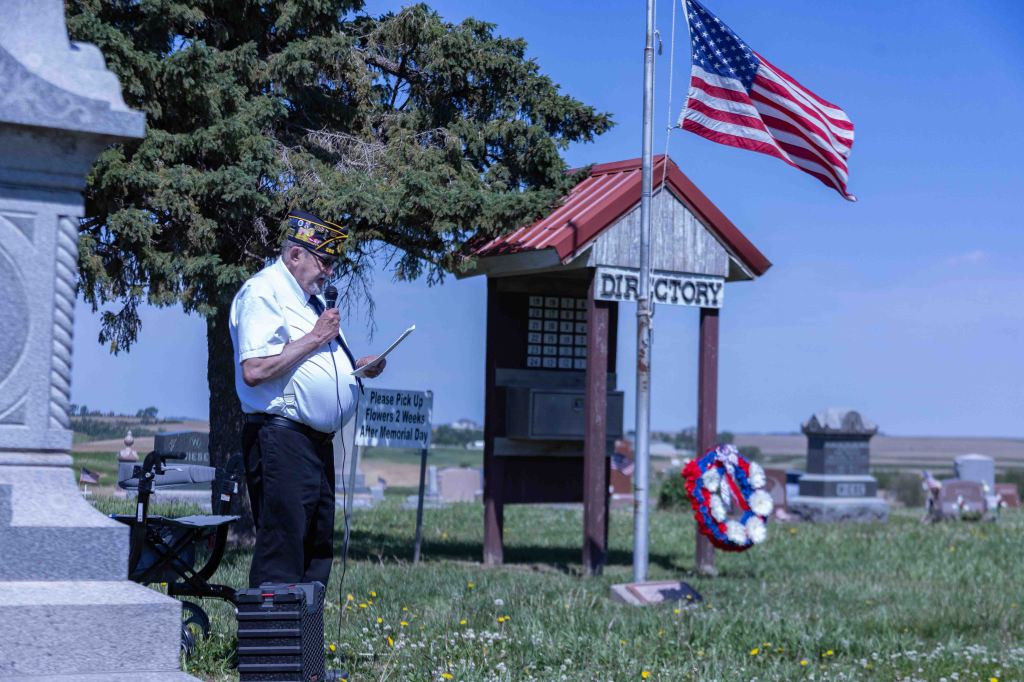 A veteran in a military cap speaks into a microphone while holding papers, standing near a flagpole with an American flag and a cemetery directory sign. A floral wreath is nearby, and gravestones are visible in the background.