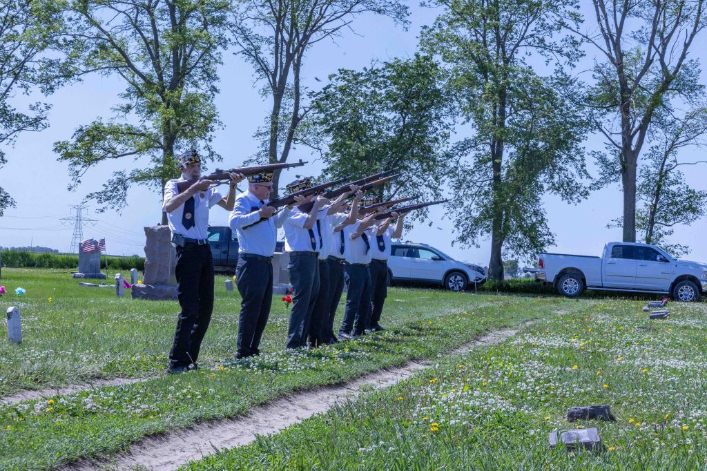 A group of uniformed individuals performs a rifle salute at a gravesite in a cemetery, surrounded by trees and flowers.