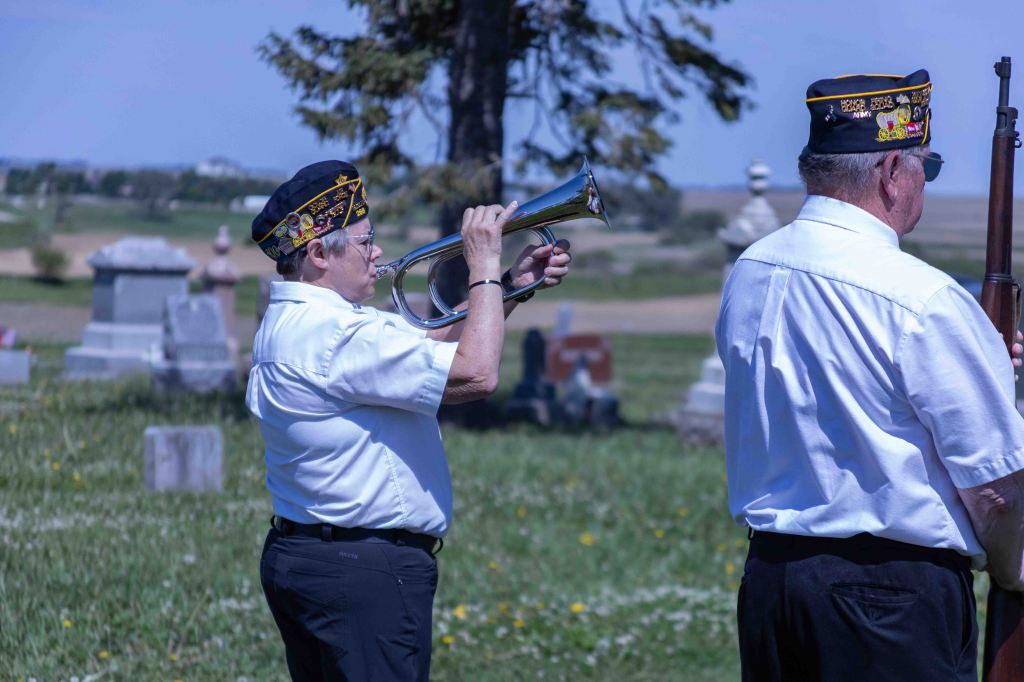 A member of a military honor guard playing the trumpet at a cemetery, while another member stands at attention nearby.