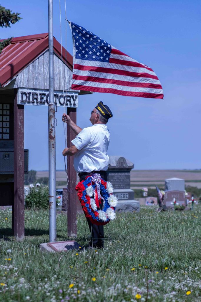 A man in a white shirt and military cap is raising an American flag at a cemetery, standing next to a directory sign. He is wearing a red, white, and blue wreath around his waist, and there are gravestones in the background.