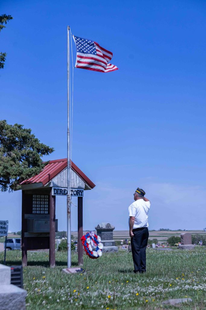A veteran stands at a cemetery looking at a waving American flag and a memorial wreath near a directory sign.