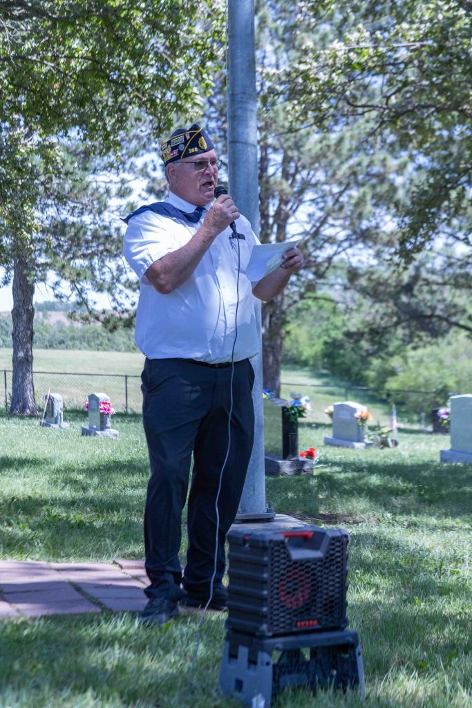 A man in a veterans' cap speaks into a microphone while holding notes, standing in a cemetery with gravestones and trees in the background.