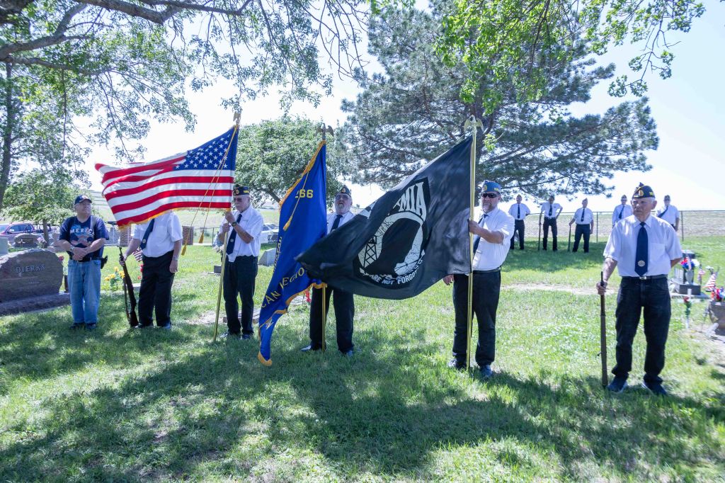 Veterans holding American and POW/MIA flags during a memorial service at a cemetery.