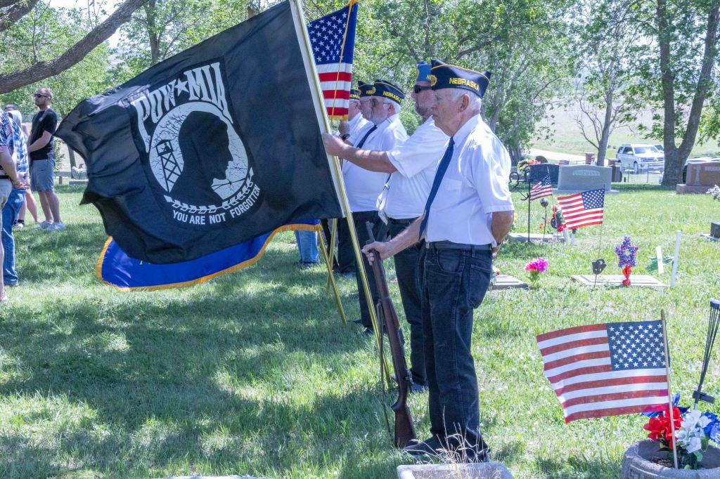 Veterans holding a POW-MIA flag and rifles at a cemetery during a memorial ceremony, surrounded by American flags and grave markers.
